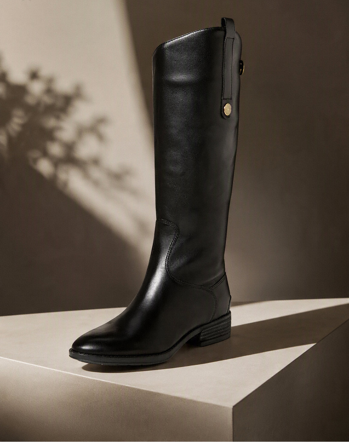 A single black leather knee-high boot with a low block heel and a small gold button detail near the top, displayed on a beige pedestal against a dark brown background with artistic leaf shadows. @ ghoddosh.com