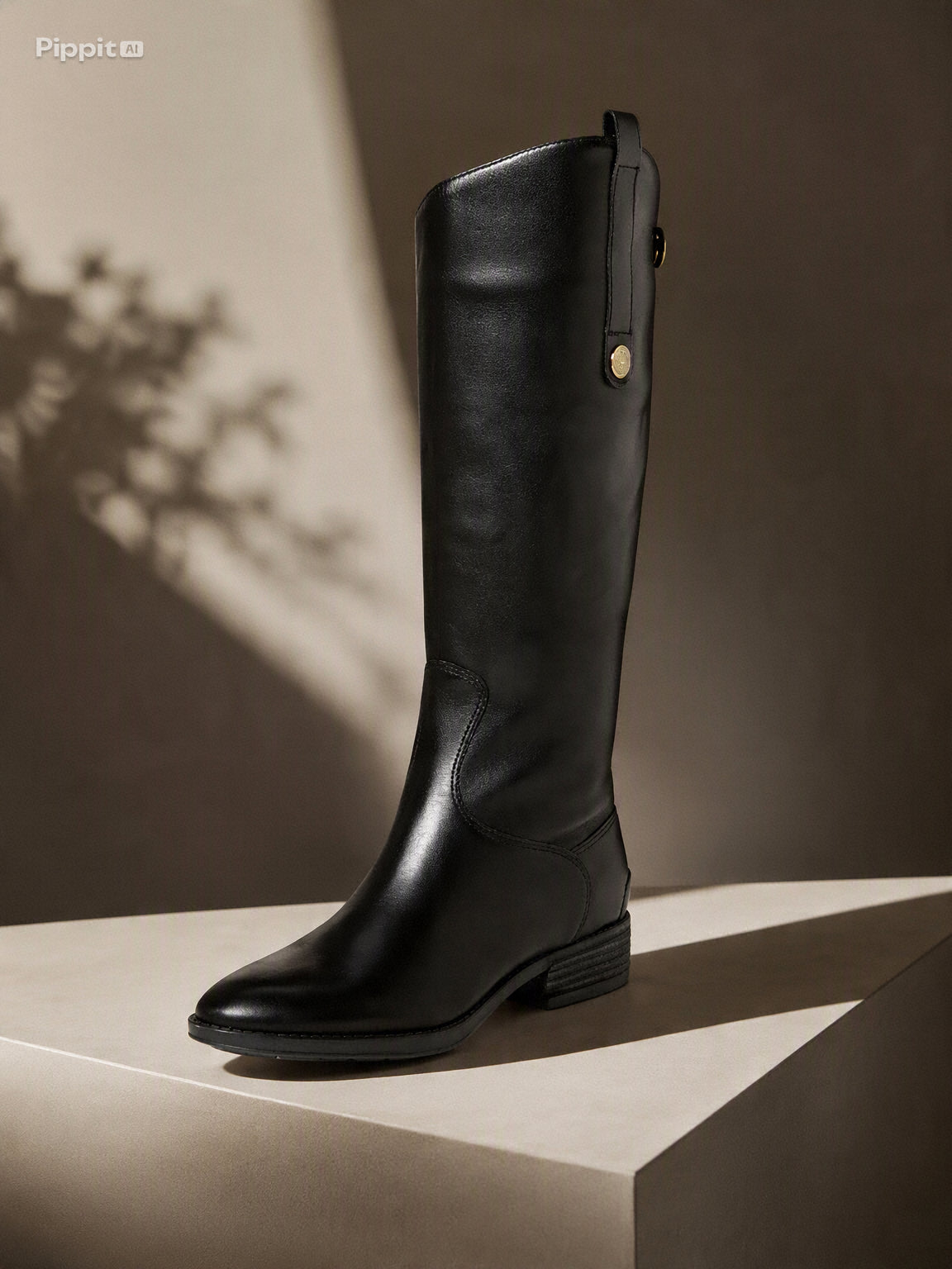 A single black leather knee-high boot with a low block heel and a small gold-tone snap detail near the top, displayed on a beige pedestal against a dark brown background with artistic leaf shadows. @ ghoddosh.com