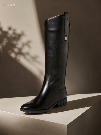 A single black leather knee-high boot with a low block heel and a small gold-tone snap detail near the top, displayed on a beige pedestal against a dark brown background with artistic leaf shadows. @ ghoddosh.com