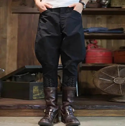 A person wearing loose black pants with decorative lace-up detailing near the lower legs, paired with dark brown boots, standing in a rustic indoor setting with wooden shelves and a fan in the background. @ ghoddosh.com