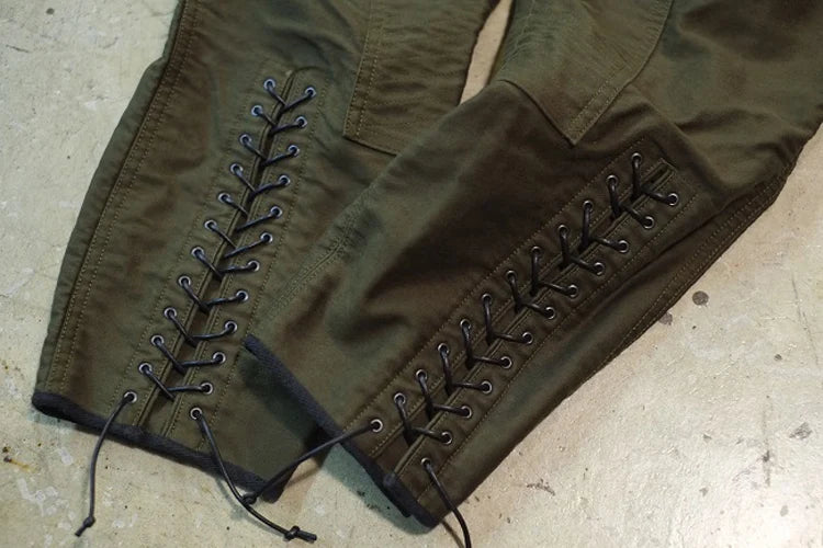 Olive green pants with black laces threaded through metal eyelets on the sides, laid flat on a concrete floor. @ ghoddosh.com
