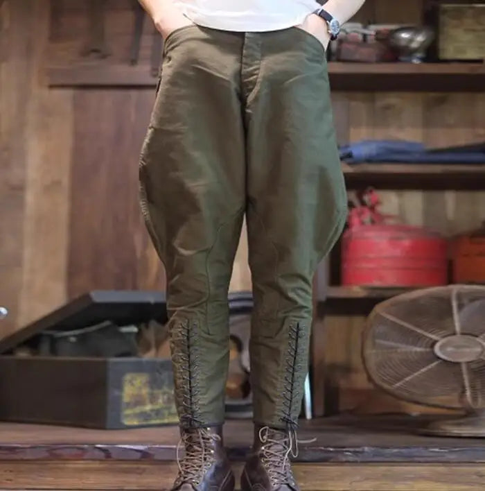 A person wearing loose olive green pants with vertical lace-up detailing down the front of the legs, paired with brown lace-up boots in a rustic indoor setting with wooden shelves and a fan in the background. @ ghoddosh.com