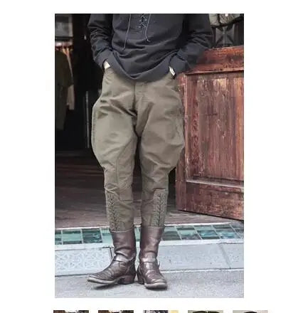 A person wearing loose olive-brown pants with decorative embroidery near the lower legs, paired with a dark hoodie and brown boots, standing in front of a rustic wooden door. @ ghoddosh.com