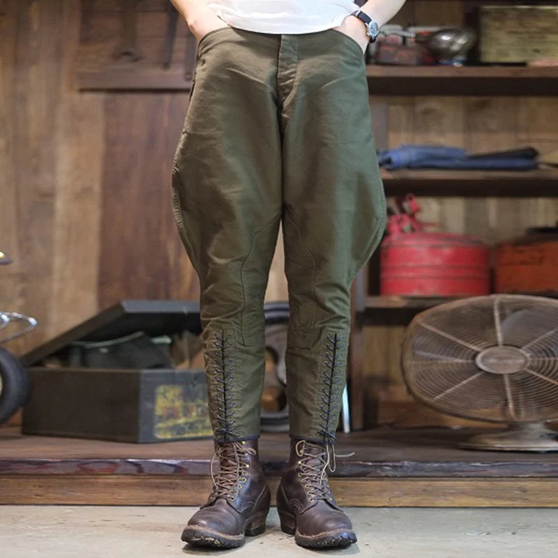 A person wearing olive green baggy pants with lace-up detailing at the ankles, paired with brown lace-up boots, standing in a rustic workshop environment. @ ghoddosh.com