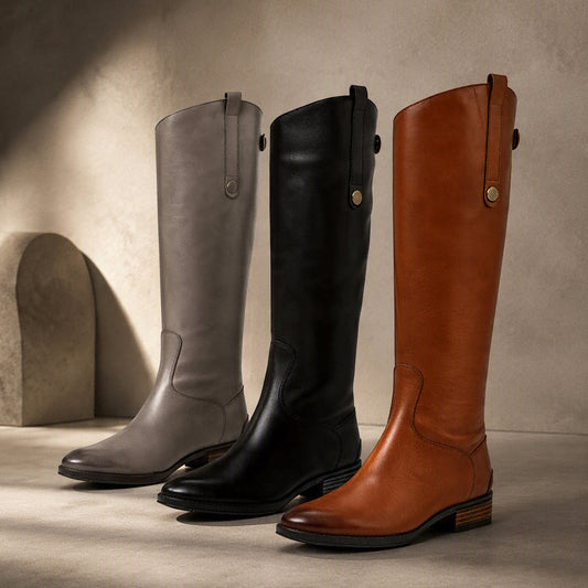 Three tall leather boots displayed side by side in a studio setting: one taupe-gray, one black, and one tan-brown. The boots are photographed against a warm beige textured wall and floor, with dramatic side lighting casting shadows. @ ghoddosh.com