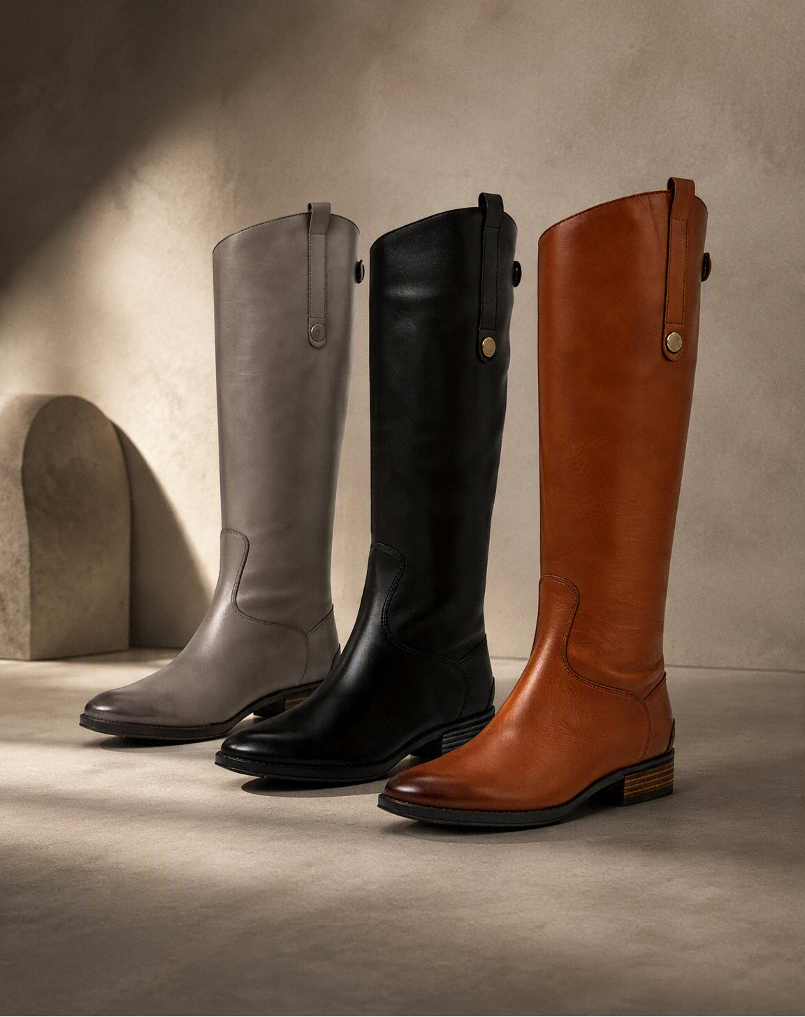 Three tall leather riding boots in gray, black, and brown, arranged in a row against a textured beige wall with dramatic side lighting. The Pippit brand logo is visible in the top left corner. @ ghoddosh.com