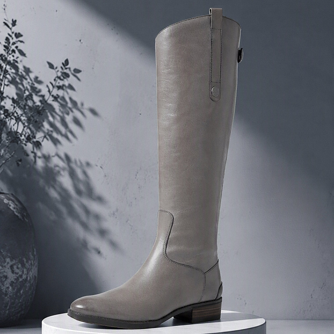 A tall light gray leather boot with a low block heel and side zipper, displayed on a white marble pedestal against a gray wall with artistic leaf shadows. @ ghoddosh.com