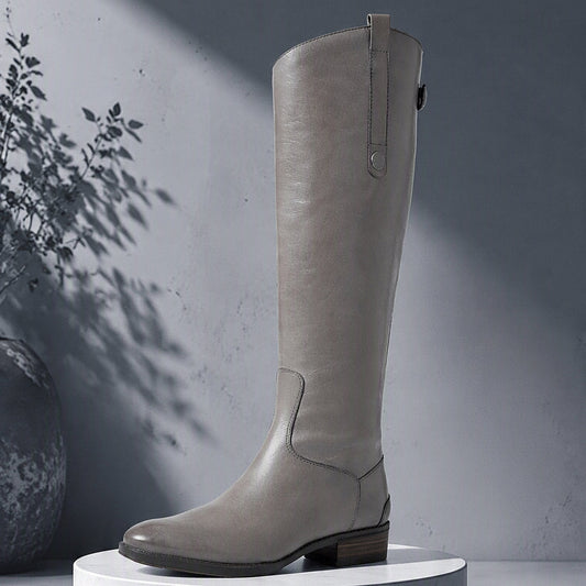 A tall light gray leather boot with a low block heel and side zipper, displayed on a white marble pedestal against a gray wall with artistic leaf shadows. @ ghoddosh.com