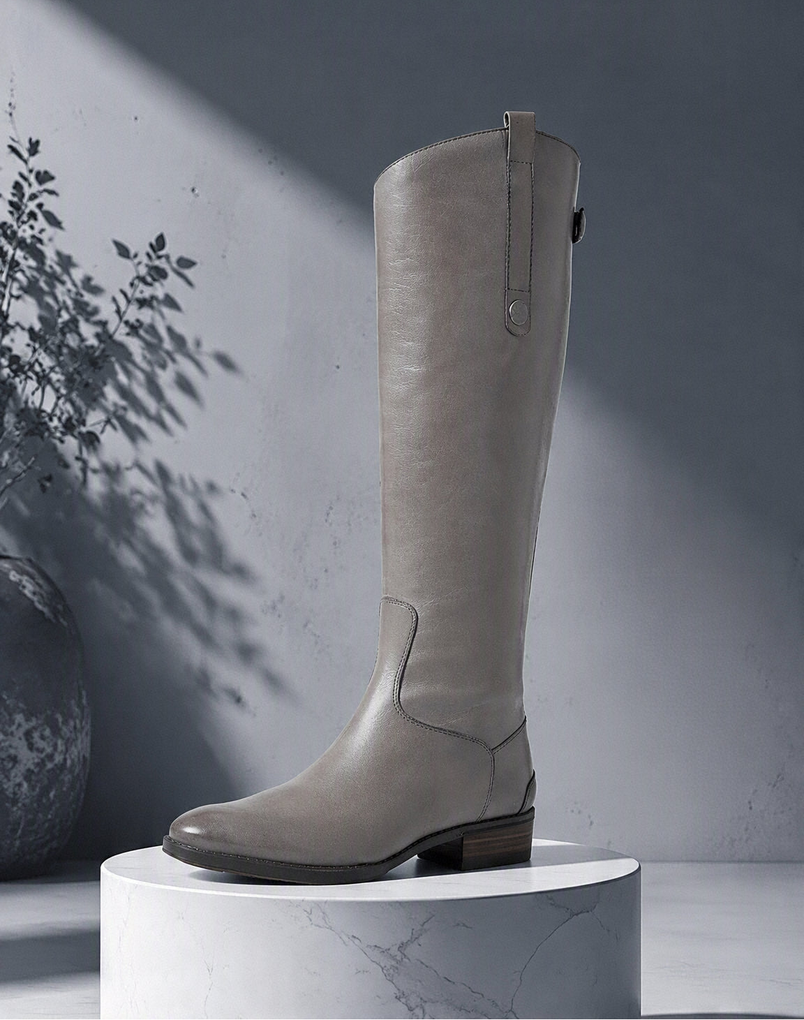 A tall taupe-brown leather knee-high boot with a low block heel and side zipper, displayed on a white marble pedestal against a gray wall with artistic leaf shadows. @ ghoddosh.com